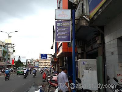 ホーチミンの大通り沿いの街角。バイクが行き交い、店先に青い案内看板が立つ風景