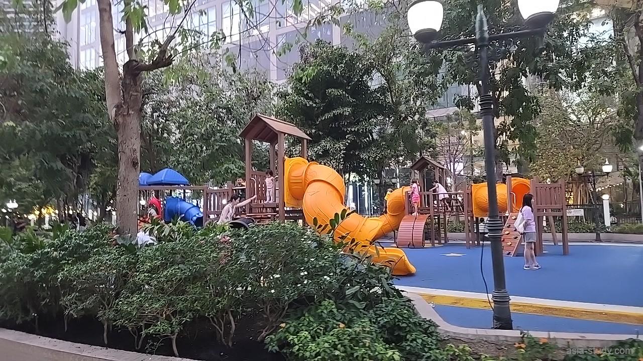 オレンジ色のスライダーや青い遊具が設置された子供用プレイエリア、木々に囲まれた家族連れで賑わう午後の公園