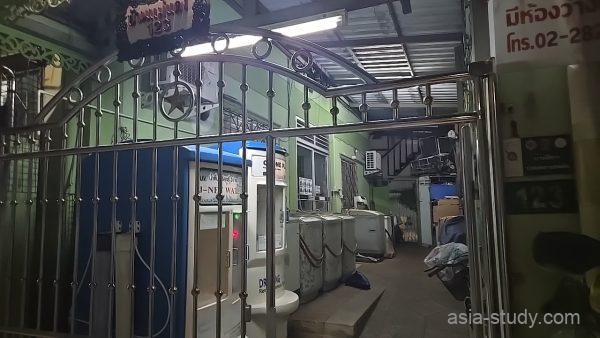 金属製の門扉の奥に洗濯機と水販売機が並ぶ、バンコクのローカルなアパートの入り口