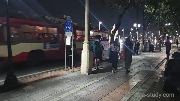 夜のバス停、走り抜ける赤いバスとバックパックを背負って待つ旅行者たち