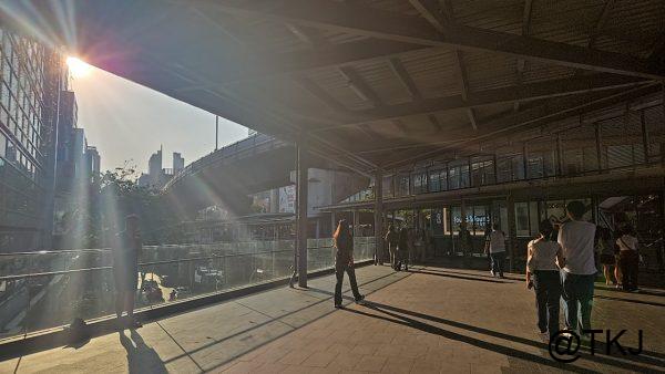 夕方のBTS駅前通路、逆光で伸びる影と行き交う人々