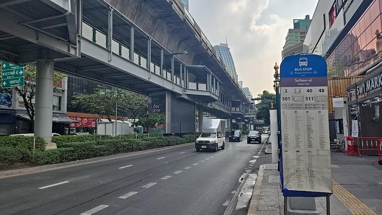 高架下のバス停ポールと路線案内、車道を走る車と夕方の空（バンコク）