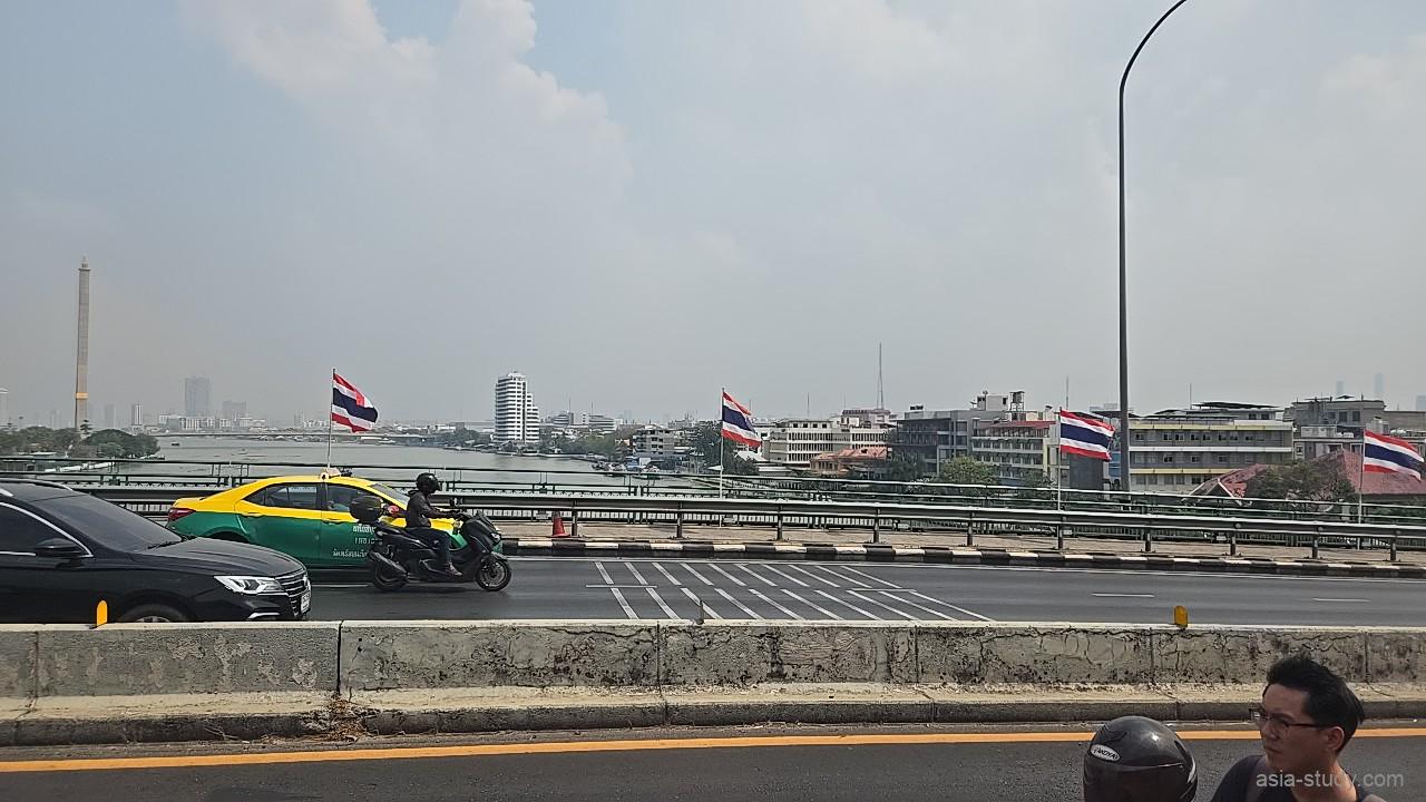 橋の上から見た川沿いの景色とタイ国旗、対岸に低層ビルが並ぶ（バンコク）