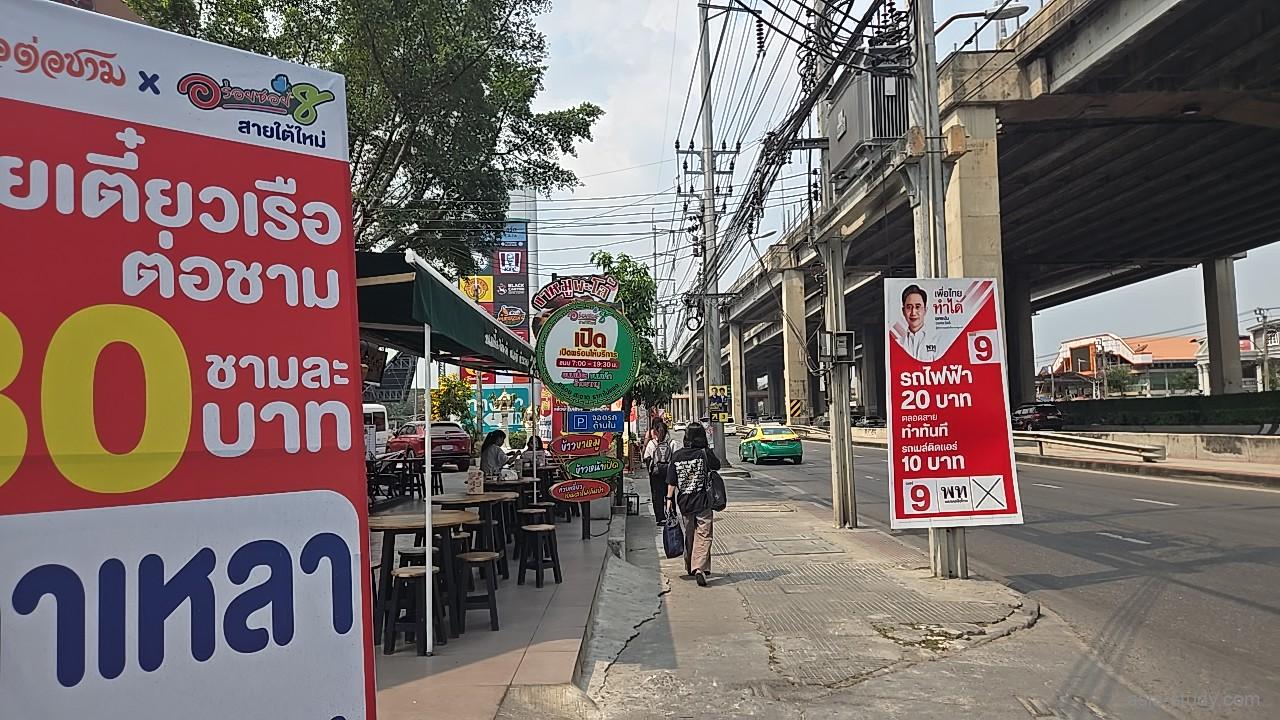 高架沿いの歩道と屋台、タイ語の大きな看板が目立つローカルな通り