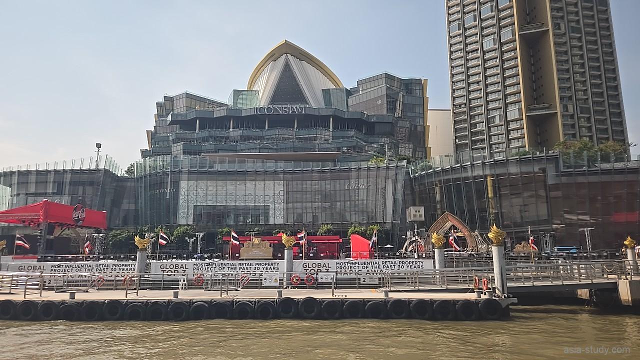 川沿いにそびえ立つアイコンサイアムのモダンな外観と、多くの旗が掲げられた専用の船着き場