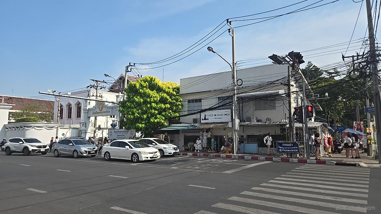 白いアーチ状の門がある寺院の入り口付近、信号待ちの車と行き交う人々が並ぶバンコクの街角