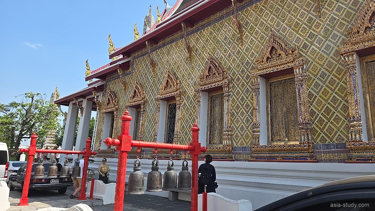 赤いフレームに吊るされた多数の鐘の列。背後の建物には豪華な金色の窓枠が見える