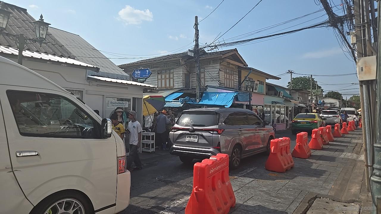オレンジ色のプラスチック製バリケードが並ぶワット・アルン裏手の通り、白いワゴン車やタイの伝統衣装レンタルショップの看板が見える街並み