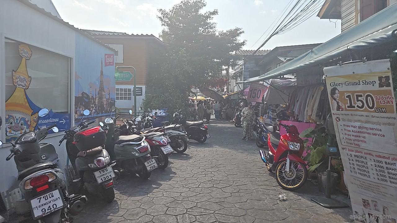パークされたバイクが並ぶ石畳の細い路地、右側には「Glory Grand Shop」などタイの伝統衣装レンタル店が軒を連ねる風景