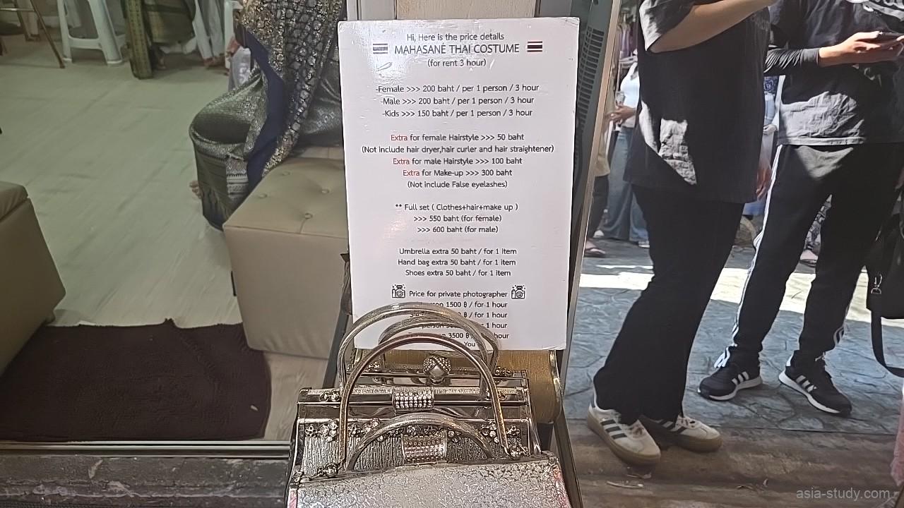 シルバーの煌びやかなハンドバッグが手前に並ぶタイ衣装レンタル店の店頭、背景にはフルセットのレンタル料金などが記された価格表