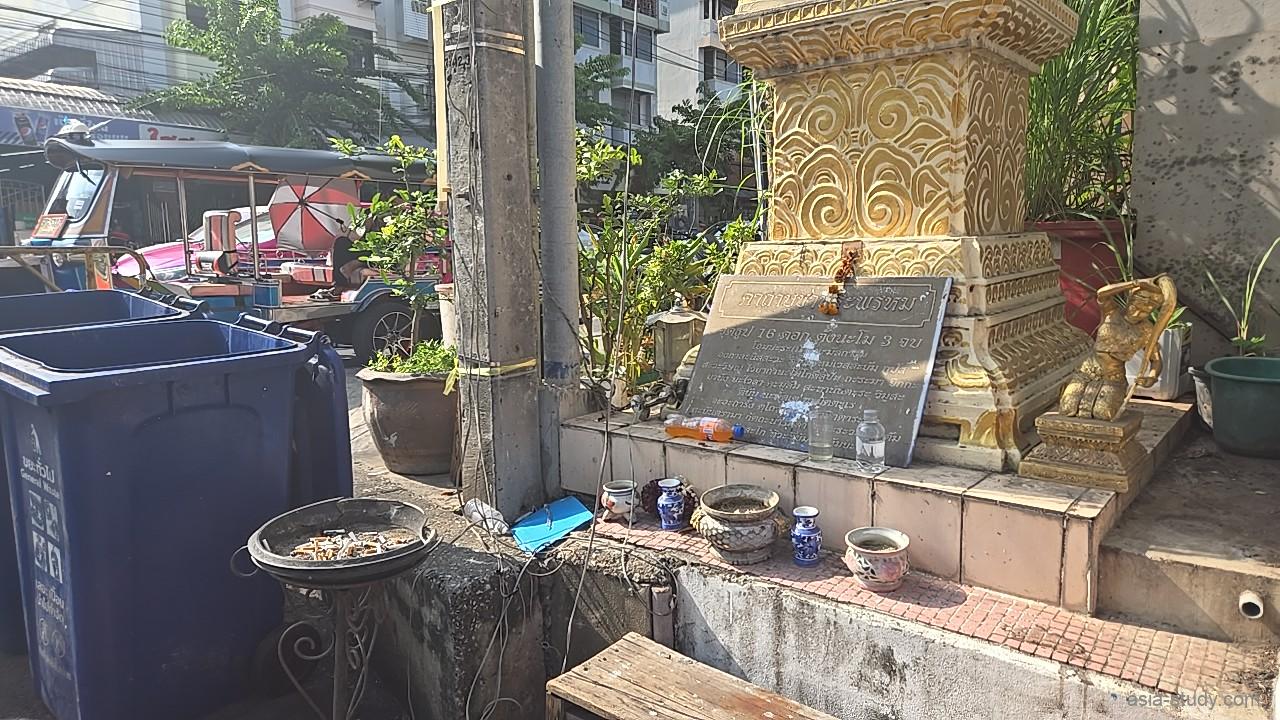 街角にある金色の装飾が施された小さな祠（ほこら）、お供え物の水や線香が置かれ、隣には青いゴミ箱とトゥクトゥクが見える風景