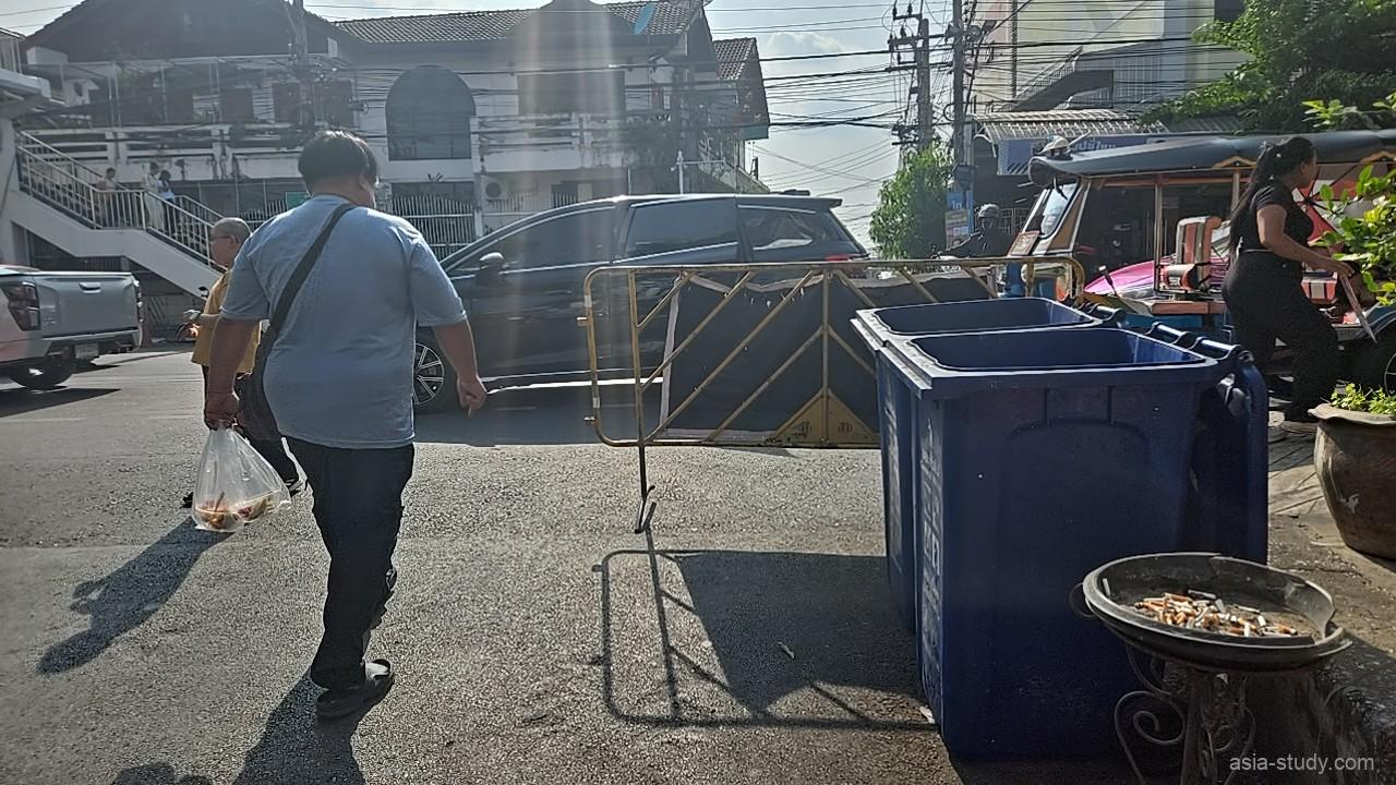 バンコクの街角の交差点、青いゴミ箱や金属製のバリケードが置かれ、買い物袋を持った通行人が歩く日常の風景