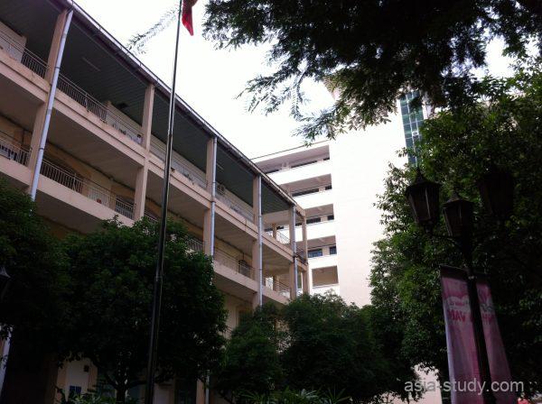 校舎の中庭に立つ旗竿と、ベージュ色の校舎（木々に囲まれたキャンパス風景）