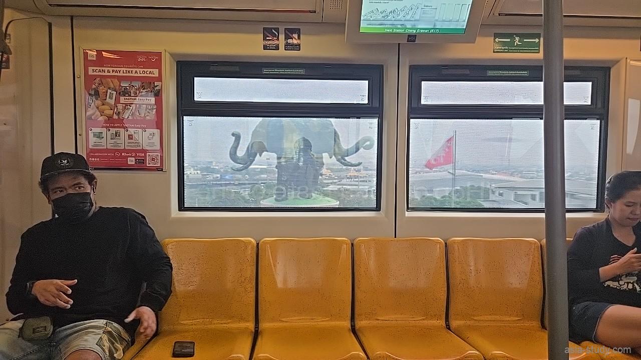 車内の黄色い座席と乗客、窓越しに見えるエラワン・ミュージアムの巨大象